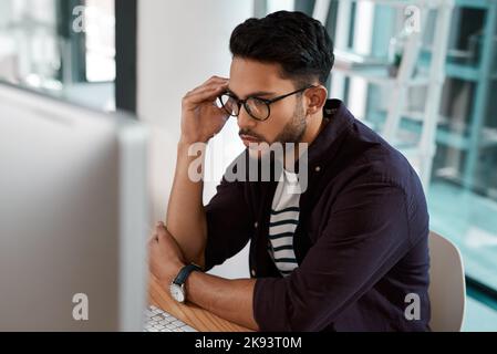 C'est intéressant. Un beau jeune homme d'affaires assis seul et regardant contemplatif tout en utilisant son ordinateur dans le bureau. Banque D'Images