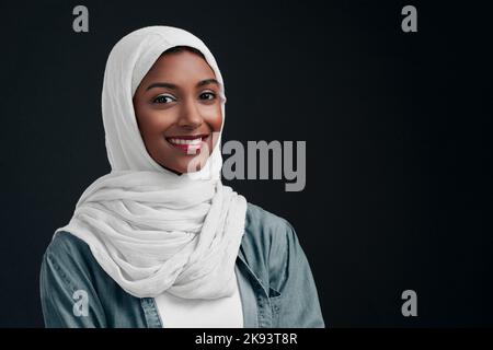 Porter mon hijab et briller avec fierté. Une jeune femme attrayante portant un hijab et debout seule contre un fond noir dans le studio. Banque D'Images