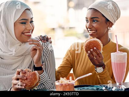 Les gourmets savent comment se rendre heureux. Deux jeunes amies affectueuses mangeant des hamburgers dans un café tout en étant vêtues de hijab. Banque D'Images