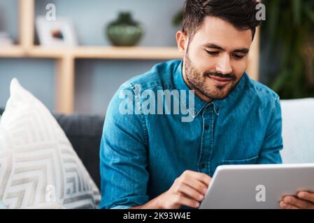 Les médias sociaux sont éclairés aujourd'hui. Un jeune homme charmant qui a l'air gai en utilisant une tablette numérique dans son salon à la maison. Banque D'Images