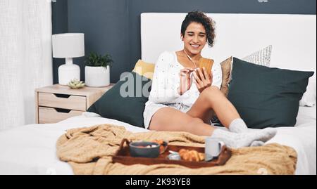 Vérification de ce qui se trouve sur les réseaux sociaux. Prise de vue en longueur d'une jeune femme attrayante écoutant de la musique depuis son téléphone portable tout en prenant le petit déjeuner au lit. Banque D'Images