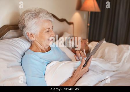 En regardant le profil en ligne de mes filles, une femme âgée assise au lit et utilisant une tablette pendant que son mari dort à côté d'elle. Banque D'Images