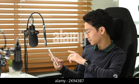 Jeune homme asiatique radio hôte assis devant le microphone à condensateur et à l'aide d'un téléphone mobile Banque D'Images