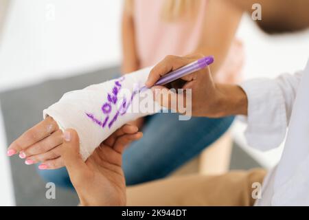 Gros plan court de mâle méconnaissable drôle de médecin écrivant l'inscription de plaisanterie sur le casting de femme avec la main cassée enveloppée dans le bandage de plâtre. Banque D'Images