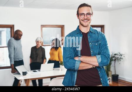 Je suis heureux que j'ai décidé de travailler ici. Portrait d'un jeune homme d'affaires gai debout avec ses bras pliés et ses collègues de travail dans le Banque D'Images