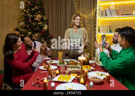 Fête du nouvel an, un groupe de personnes de divers amis fêtent Noël ensemble, hommes et femmes se réjouissent et mangent à la table, buvez du vin, prenez des photos de nouveaux plats et la vidéo en ligne. Banque D'Images