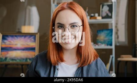 Gros plan heureuse femme funy avec les cheveux rouges peintre fille artiste en lunettes regarde la caméra étire les mains vers l'avant montrant les paumes sales tachées dans l'acrylique Banque D'Images