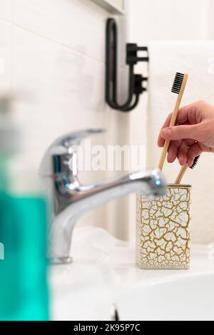 La main d'une femme place une brosse à dents en bois dans un verre avec un motif or dans la salle de bains sur fond d'un robinet et de carreaux brillants.Sélectif Banque D'Images