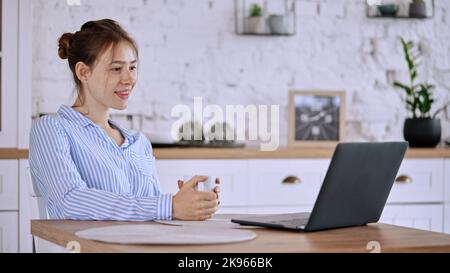 Une jeune femme dans une chemise bleue rayée buvant du thé lors d'un appel vidéo avec un ordinateur portable Banque D'Images