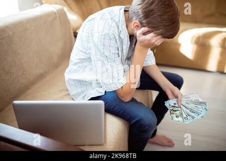 Triste adolescent regardant un paquet de dollars dans les mains tout en étant assis à côté d'un ordinateur portable à la maison. Problèmes de dépôts d'argent et d'épargne chez les adolescents. Tôt Banque D'Images