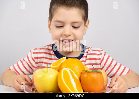 Un garçon d'âge préscolaire regarde les fruits - oranges, persimmons et pommes sur un fond blanc. Banque D'Images