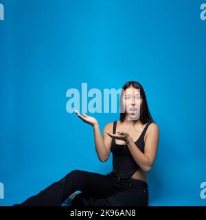 Jeune multiracial tendance surpris femme assise tenant et présentant l'espace de copie sur sa paume isolée sur fond bleu Banque D'Images