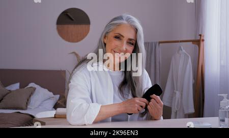 Portrait personne âgée adulte femme d'âge moyen âgée de 50s ans femme caucasienne en peignoir matin beauté routine regardant l'appareil photo peignant gris long brillant Banque D'Images