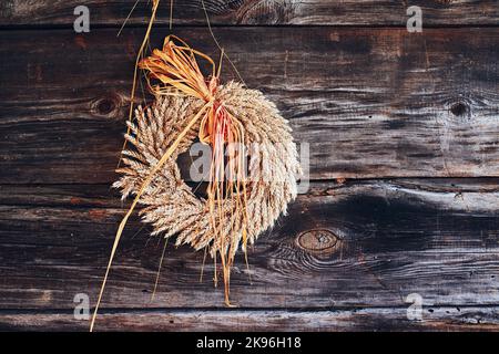 Couronne d'oreilles dorées de blé, fleurs séchées et herbes attachées avec un ruban rouge accroché sur un mur en bois. Décoration rustique naturelle Banque D'Images