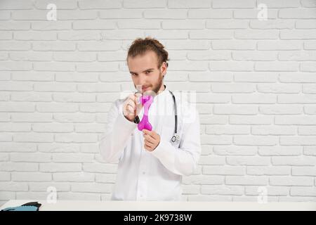 Portrait de vétérinaire masculin en robe blanche avec cheveux bouclés dans le bandeau montrant le jouet de chien rose en caoutchouc pour mâcher pendant la dentition. Véritable spécialiste toujours prêt à aider nos petits frères - animaux. Banque D'Images