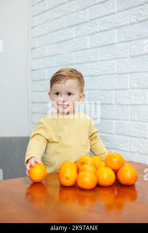 Un petit garçon de 2 ans est debout à une table avec des mandarines. Bébé veut s'asseoir sur les agrumes pour la première fois Banque D'Images