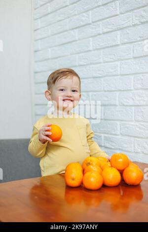 Un petit garçon de 2 ans est debout à une table avec des mandarines. Bébé veut s'asseoir sur les agrumes pour la première fois Banque D'Images