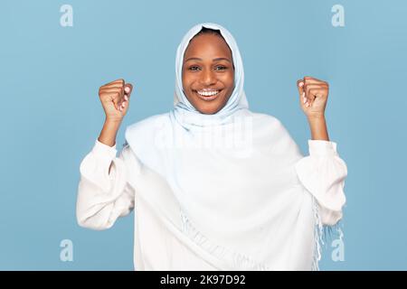 Femme musulmane noire et émotive dans le hijab célébrant le succès, accrochant les poings et souriant sur fond bleu de studio Banque D'Images