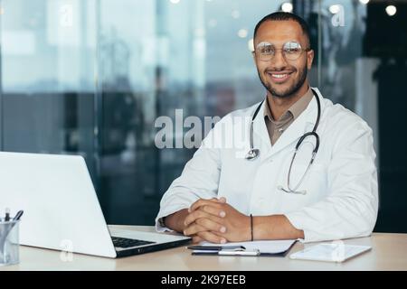 Portrait du jeune médecin pédiatre hispanique masculin. Assis dans le bureau à une table avec des documents et un ordinateur portable, portant des lunettes et un stéthoscope dans un manteau blanc, regardant la caméra, souriant. Banque D'Images