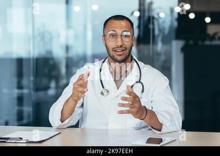 Un jeune médecin latino-américain est assis dans le bureau face à la caméra et mène une consultation en ligne avec un patient, organise un séminaire en ligne médical. Il parle, explique, fait des vagues. Banque D'Images