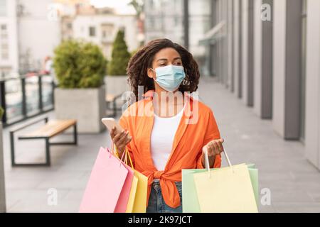 Bonne jeune femme afro-américaine en décontracté, masque de protection avec beaucoup de sacs et smartphone Banque D'Images