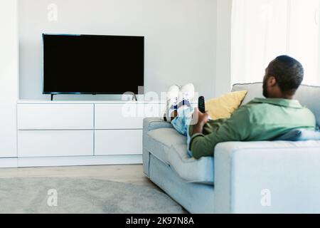 Homme noir regardant la télévision avec écran vierge, en tenant la télécommande et en changeant de chaîne sur écran plat plasma, maquette Banque D'Images