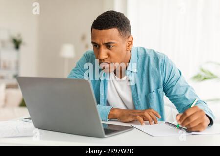 Sérieux concentré s'ennuyer jeune africain américain mâle dans les jeans regardant l'ordinateur portable faire des notes, trouver erreur Banque D'Images