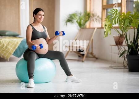 Entraînement de femme enceinte souriante avec haltères et assis sur Fitball à la maison Banque D'Images