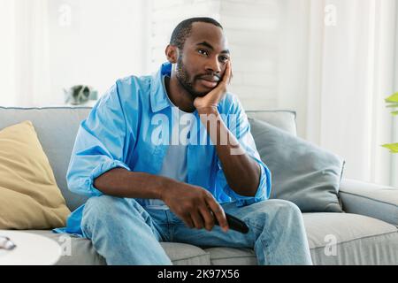 Programme de télévision ennuyeux. Un homme noir décontentant regardant la télévision, changeant de chaîne avec télécommande, assis sur un canapé Banque D'Images