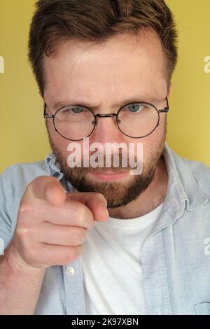 Homme avec une barbe et des lunettes regarde et pointe strictement les doigts, sur un fond jaune. Banque D'Images