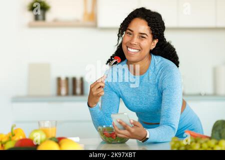 Bonne dame noire dans les vêtements de sport manger de la salade à la maison dans la cuisine, se pencher sur la table et en utilisant le smartphone, l'espace libre Banque D'Images