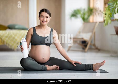 Femme enceinte souriante en vêtements d'activité posant sur un tapis de yoga à la maison Banque D'Images