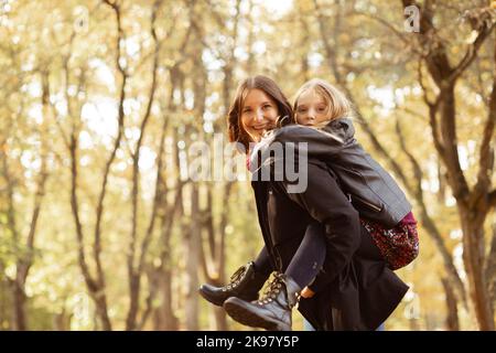 Une jeune femme joyeuse se balade dans la vue latérale de la petite fille à l'arrière. Bonne mère et fille dans des tenues noires décontractées ont le plaisir et jouent dans le parc d'automne doré. Automne Banque D'Images