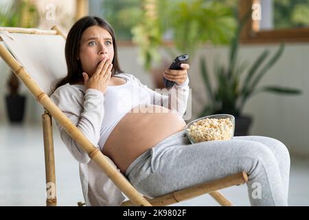 Contenu du choc. Femme enceinte effrayée regardant la télévision et Eating Popcorn à la maison Banque D'Images