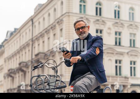 Homme mûr à poil gris moderne utilisant son smartphone et à vélo dans les rues de la ville avec de beaux bâtiments en arrière-plan. Photo de haute qualité Banque D'Images