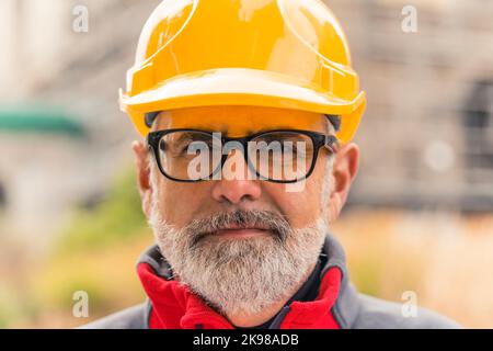 Portrait rapproché d'un ouvrier de construction professionnel d'âge moyen, confiant, caucasien, barbu, aux cheveux gris, portant en uniforme un casque orange vif et des lunettes, regardant la caméra. Photo de haute qualité Banque D'Images