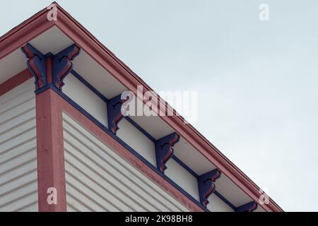 Une section d'angle de toit plat d'un grand bâtiment blanc en bois d'époque avec garniture décorative en bois rose et violet sous l'éve. La garniture est rose Banque D'Images