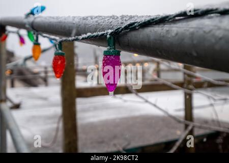 Une série de lumières de Noël colorées entourant une main courante en métal noir. Il y a de la neige blanche et fraîche sur le rail et le sol en béton. Banque D'Images