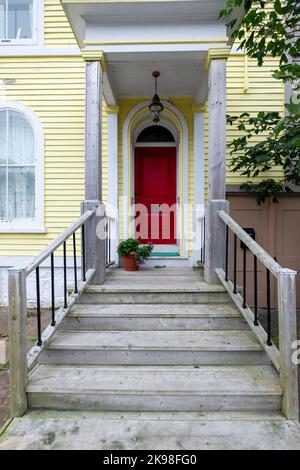 L'extérieur d'une maison jaune vif avec garniture blanche. Il y a plusieurs fenêtres et une porte rouge vibrante avec des marches en bois menant à l'entrée Banque D'Images