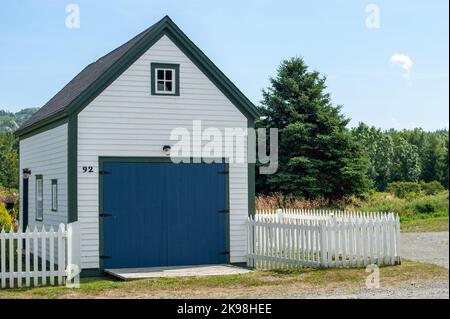 Un garage ou une grange en bois blanc de deux étages avec une porte de garage bleu profond et plusieurs petites fenêtres en verre. Il y a une clôture de piquetage blanche qui enferme une cour Banque D'Images