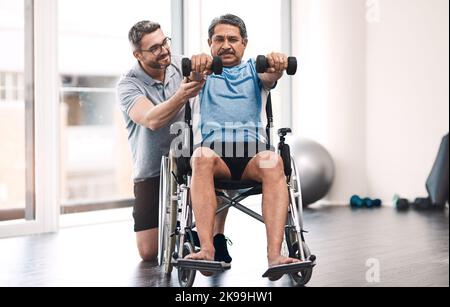 Muscle de construction. Plein coup d'un homme âgé dans un fauteuil roulant s'exerçant avec des haltères le long de son physiothérapeute. Banque D'Images