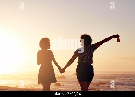 Les amis sont des gens qui aiment la vie ensemble. Vue arrière de deux jeunes femmes méconnaissables se tenant les mains et marchant le long de la plage au coucher du soleil. Banque D'Images