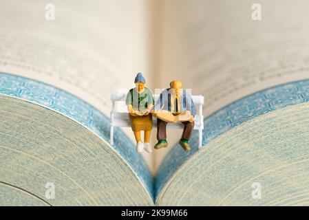 Gros plan de figurines miniatures d'un couple âgé lisant un livre et un journal sur un banc. Banque D'Images