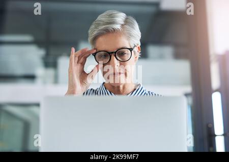 En regardant de plus près les choses, une femme d'affaires attirante et mûre qui ajuste ses lunettes tout en travaillant sur un ordinateur portable dans son bureau. Banque D'Images