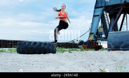 Une jeune femme sportive dans un haut rouge et des leggings noirs effectue différents exercices de force en utilisant une grande roue de tracteur lourde, forme ses muscles. Derrière est port de cargaison, l'aube. Photo de haute qualité Banque D'Images