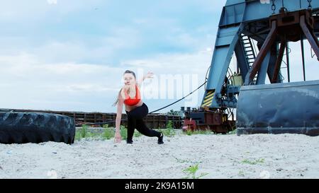 Une jeune femme sportive dans un haut rouge et des leggings noirs effectue différents exercices de force en utilisant une grande roue de tracteur lourde, forme ses muscles. Derrière est port de cargaison, l'aube. Photo de haute qualité Banque D'Images