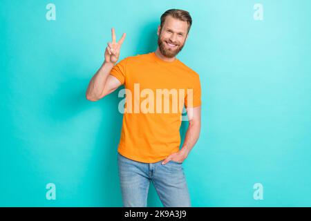 Portrait d'un homme agréable et confiant avec une barbe blonde porter un t-shirt orange main dans la poche montrant le V-signe isolé sur fond de couleur sarcelle Banque D'Images