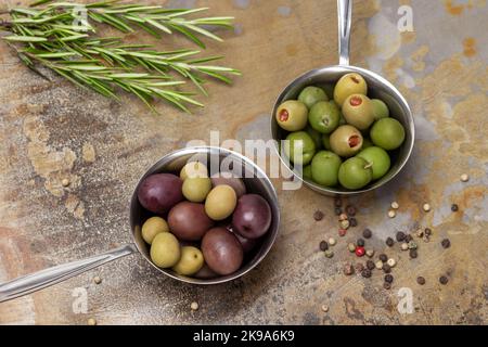 Olives de différentes variétés dans un bol en métal. Romarin et allépice. Pose à plat. Arrière-plan en métal rouillé. Banque D'Images