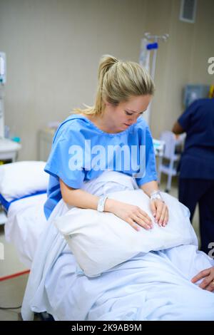 Je prie tout va selon le plan. Une jeune femme enceinte assise sur son lit d'hôpital et regardant réfléchie avec des infirmières en arrière-plan. Banque D'Images