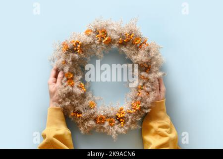 Femme tenant dans les mains couronne de Thanksgiving avec des fleurs orange et des matériaux naturels secs sur fond bleu. Vue de dessus. Banque D'Images